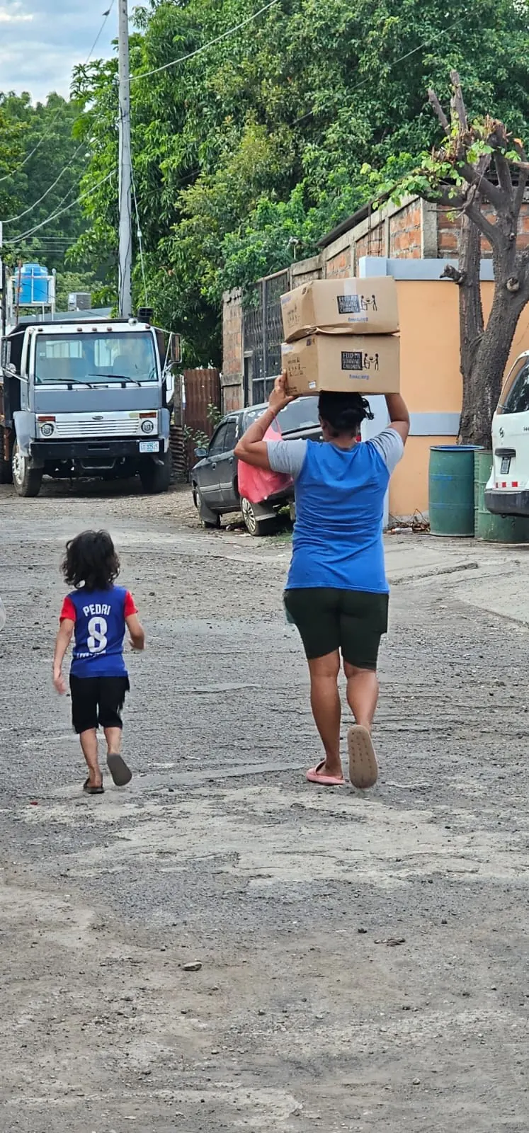 A woman carrying feeding-program supply boxes with a child walking beside her